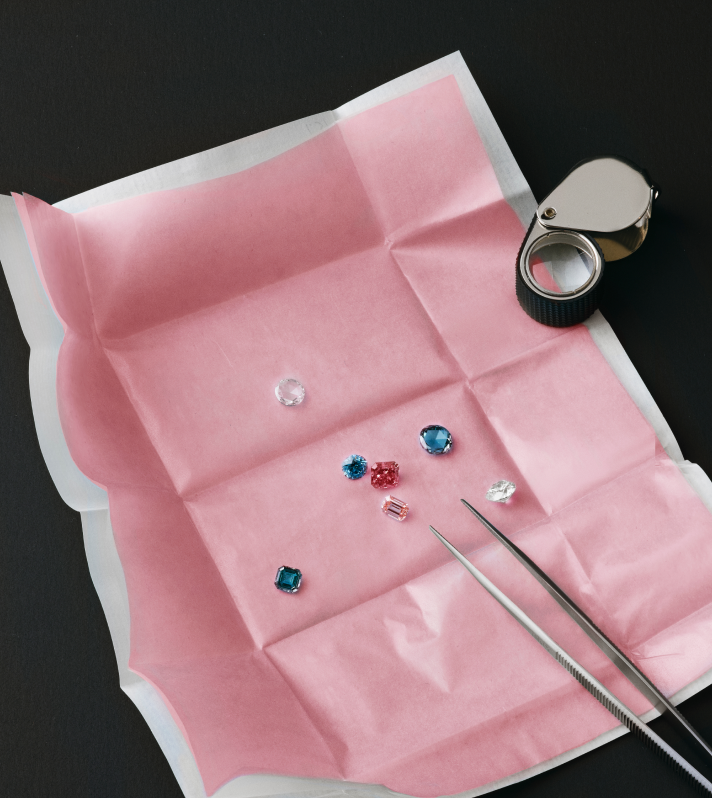 Pierres précieuses colorées sur papier rose avec pince joaillier et loupe d’observation.
